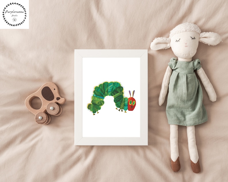 Op de afbeelding: Een wit ingelijst afdruk van een groene rups met rode ogen en een rode mond. De rups bevindt zich op een witte achtergrond. De afdruk is op een beige bedsprei met een knuffeldier-schaap en een houten speelgoed.