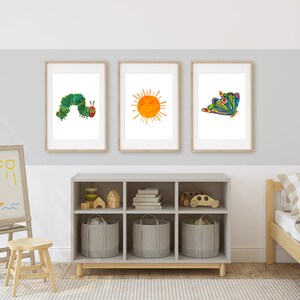 Op de afbeelding: Drie ingelijste prints op een witte muur. De prints tonen een groene rups, een gele zon en een kleurrijke vlinder. De prints bevinden zich in een kinderkamer met een houten ezel, een boekenkast en een bed.
