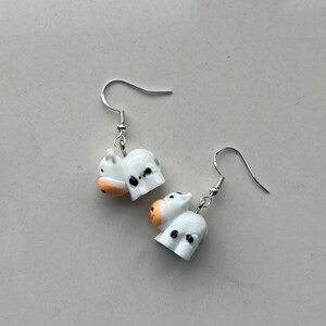 May include: A pair of white cow-shaped earrings with black spots and orange muzzles, hanging from silver-coloured fish hook ear wires. The cow earrings are a fun and whimsical accessory.