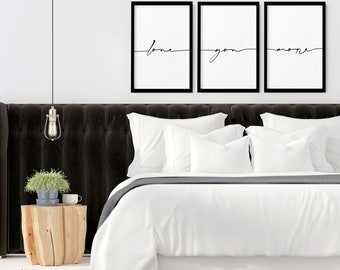 May include: Three rectangular wall art prints in black frames featuring the cursive text love, you, more. The prints are displayed above a bed with a dark tufted headboard, white bedding, and a light wood side table with a small green plant.