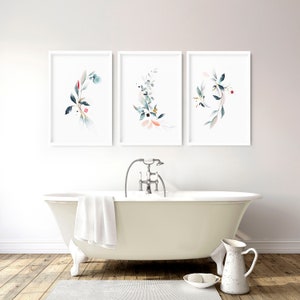 May include: Three framed wall art prints featuring watercolor botanical illustrations of leaves and berries in shades of blue, green, and soft pink. Each print is displayed in a simple white frame, arranged horizontally above a bathtub.