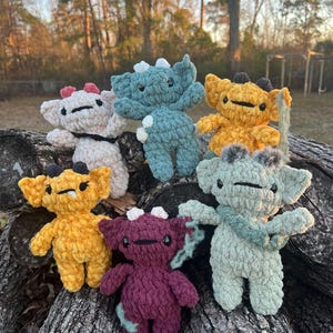 May include: A collection of handmade crochet monster dolls in various colors, including yellow, purple, and teal. Each doll has a unique design with small horns and expressive faces. The dolls are displayed on a weathered wooden surface.