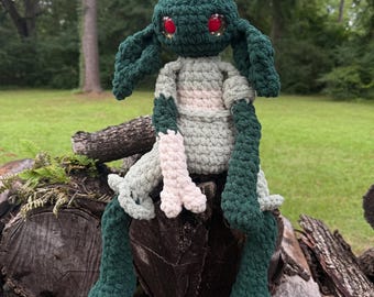 May include: A handmade crochet plush creature featuring a dark green head with large red eyes, long floppy ears, and a small white tuft on top. The body is a mix of light green and dark green yarn with long limbs and white-tipped hands and feet.