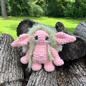 Peut inclure: Une créature en peluche crochetée à la main avec un corps rose doux, de grandes oreilles pointues et de longs cheveux en fil vert texturé. La figurine présente de grands yeux bleus brillants avec des reflets blancs et de petites ailes blanches.