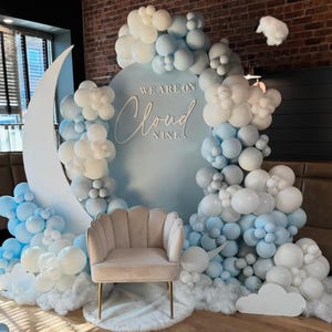 May include: A baby shower backdrop with a large white crescent moon and a blue circle with the text "We are on Cloud Nine". The backdrop is decorated with white and blue balloons and clouds.