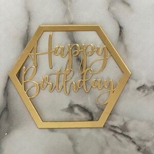 May include: A gold-colored, hexagonal cake topper with the words "Happy Birthday" in a cursive font. The topper is made of a shiny material and is set against a marble background.