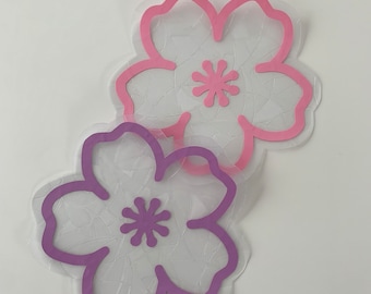 Può includere: Due sagome di fiori di carta, una rosa e una viola, con un centro bianco e uno sfondo bianco a motivi.