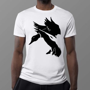 Puede incluir: Una camiseta blanca con una silueta negra de un pato volando con un ca&ntilde;&oacute;n de pistola en su pico.