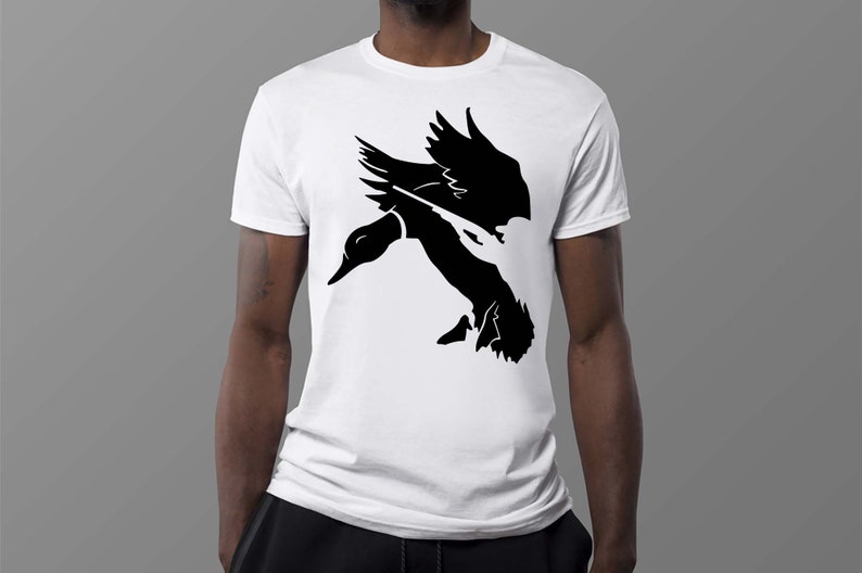Puede incluir: Una camiseta blanca con una silueta negra de un pato volando con un ca&ntilde;&oacute;n de pistola en su pico.