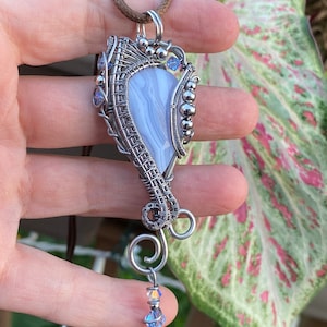May include: A silver wire wrapped pendant featuring a blue and white agate stone with a crystal accent. The pendant is attached to a brown leather cord.