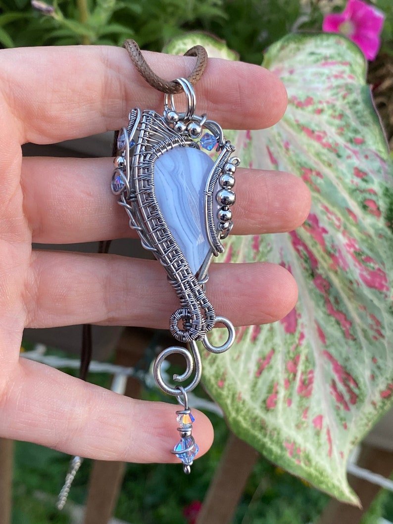 May include: A silver wire wrapped pendant featuring a blue and white agate stone with a crystal accent. The pendant is attached to a brown leather cord.