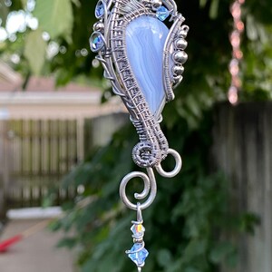 May include: A silver wire wrapped pendant featuring a light blue agate stone with a swirl pattern. The pendant has a silver chain and a small crystal dangle.