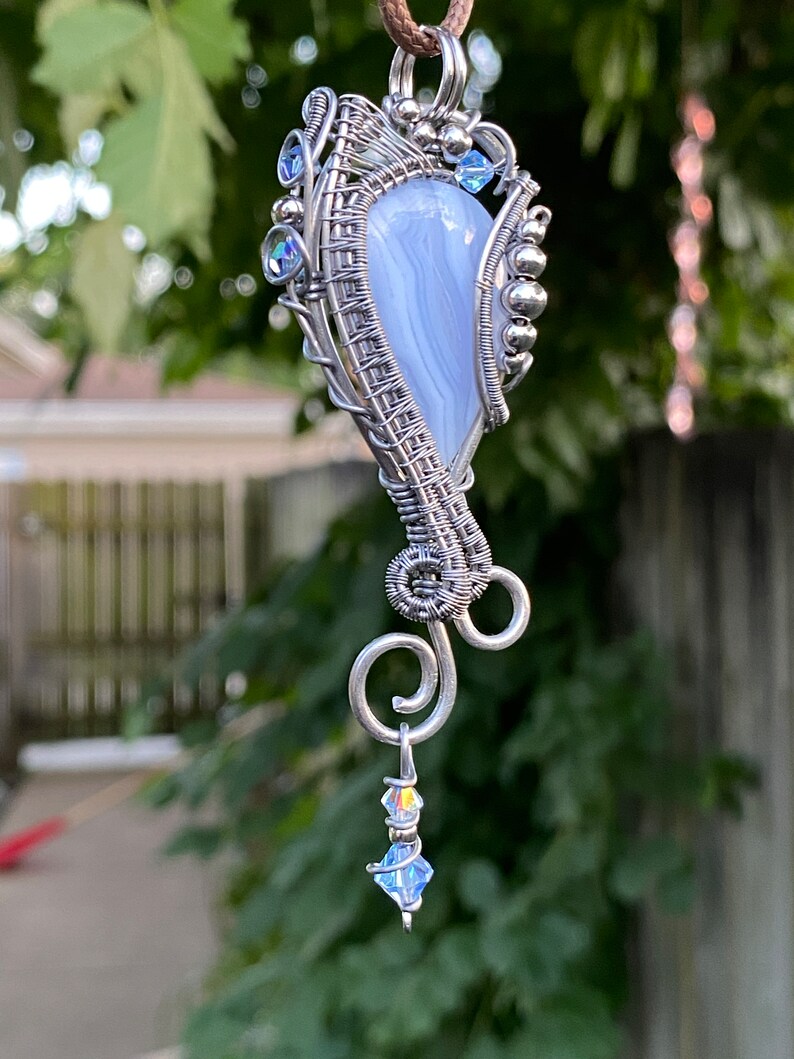 May include: A silver wire wrapped pendant featuring a light blue agate stone with a swirl pattern. The pendant has a silver chain and a small crystal dangle.