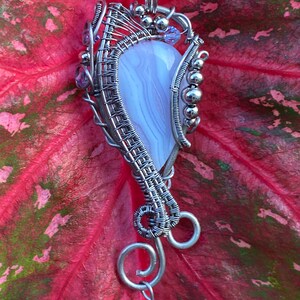 May include: A silver wire wrapped pendant featuring a light purple agate stone with a swirl pattern. The pendant is accented with small silver beads and purple crystals.
