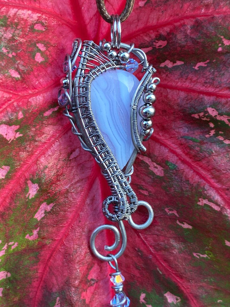 May include: A silver wire wrapped pendant featuring a light purple agate stone with a swirl pattern. The pendant is accented with small silver beads and purple crystals.