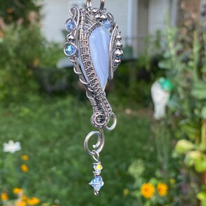 May include: A silver wire wrapped pendant featuring a light blue agate stone with a swirl pattern. The pendant has a silver chain with a small crystal bead dangling from the bottom.