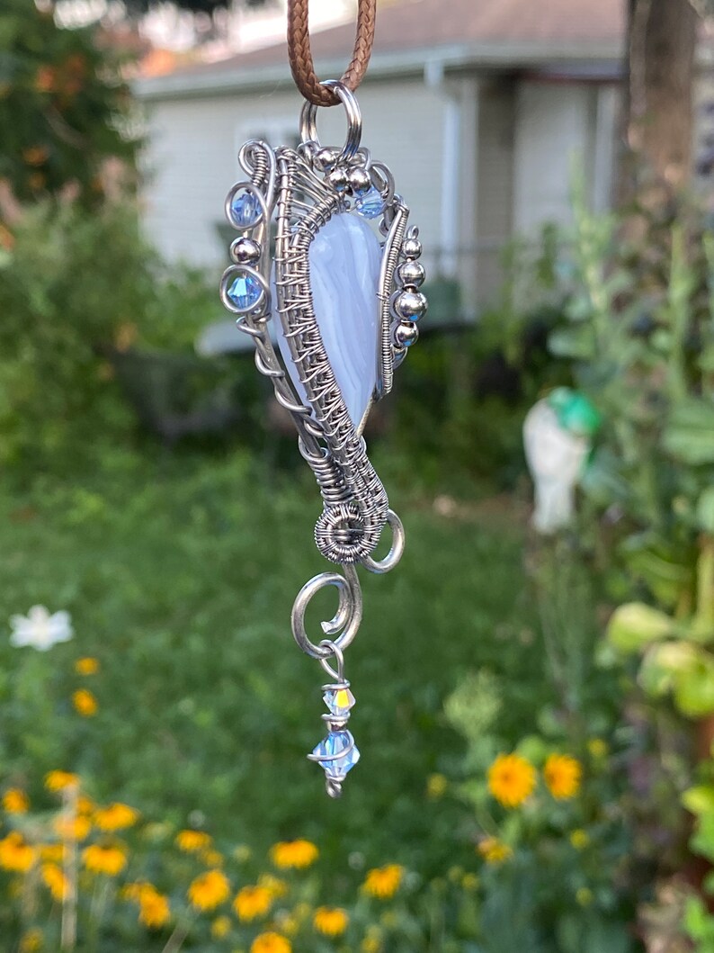 May include: A silver wire wrapped pendant featuring a light blue agate stone with a swirl pattern. The pendant has a silver chain with a small crystal bead dangling from the bottom.