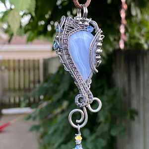 May include: A silver wire wrapped pendant featuring a blue agate stone with a swirl pattern. The pendant is accented with small clear crystals and silver beads. The pendant hangs from a brown cord.