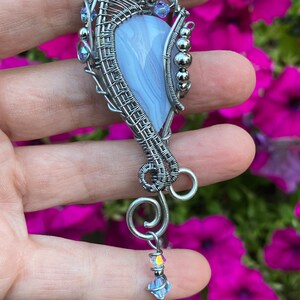 May include: A silver wire wrapped pendant featuring a light blue agate stone with a swirl pattern. The pendant is accented with small silver beads and clear crystals.