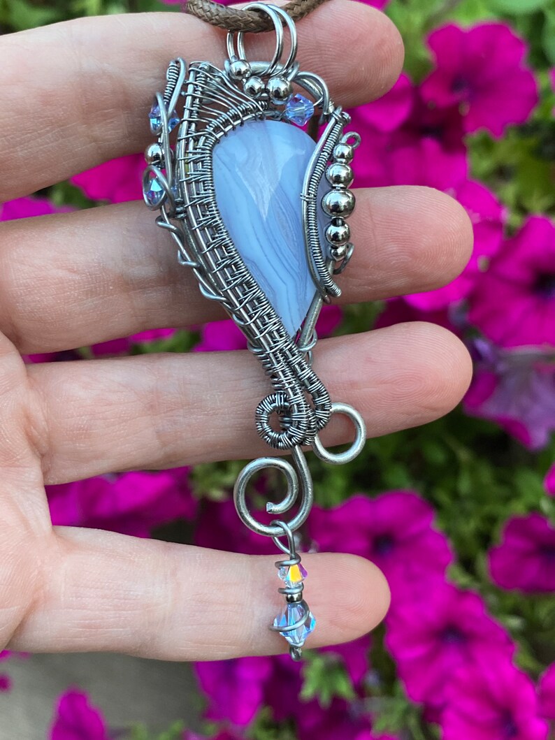 May include: A silver wire wrapped pendant featuring a light blue agate stone with a swirl pattern. The pendant is accented with small silver beads and clear crystals.