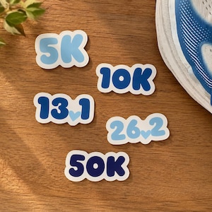 May include: A collection of five die-cut stickers featuring the text 5K, 10K, 13.1, 26.2, and 50K in shades of blue. The stickers have a white border and some include small light blue heart icons between the numbers.