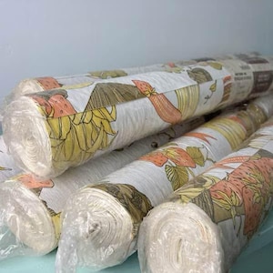 May include: Several rolls of fabric are stacked, showcasing a repeating pattern of yellow pineapples, orange strawberries, and brown leaves on a white background. The fabric is wrapped in clear plastic, ready for use.
