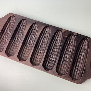 May include: Cast iron cornbread pan with six individual molds shaped like corn cobs. The pan is dark brown and has a rustic look.