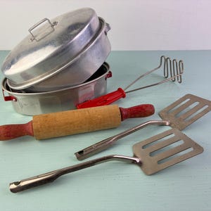 May include: A collection of vintage kitchen tools including a round aluminum pot with a lid, a potato masher with a red handle, a wooden rolling pin with red painted handles, and two metal slotted spatulas with rectangular heads and handles.