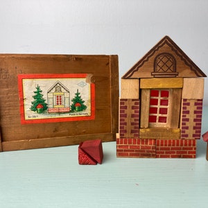 May include: Wooden toy house building blocks with a red and brown color scheme. The house features a red window, brick detailing, and a small red roof. A wooden box with a label that says "Made in Germany" is also included.