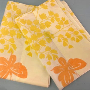 May include: Two folded vintage sheets with a floral pattern. The sheets are cream-colored with yellow and orange floral designs. The design includes yellow branches and orange flowers.