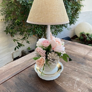 Pode incluir: Um abajur de mesa com um abajur de tecido bege e uma base de xícara de chá branca. A xícara de chá está cheia de flores rosa e brancas e uma pinha.