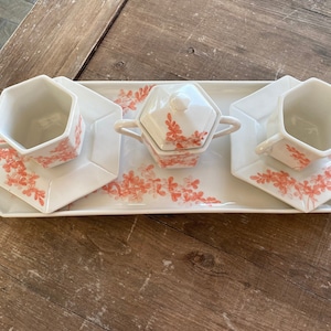 Könnte beinhalten: Ein weißes Porzellan-Tee-Set mit floralem Design. Das Set enthält eine Teekanne, zwei Tassen und zwei Untertassen. Die Tassen und Untertassen sind sechseckig. Die Teekanne hat einen Deckel mit einem Knopf in Form einer Blume.