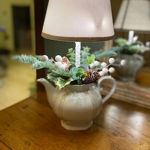 Pode incluir: Um bule branco com guarnição dourada é usado como base de lâmpada. O bule é decorado com um arranjo floral de inverno, incluindo ramos de pinho, bagas brancas e uma pinha.