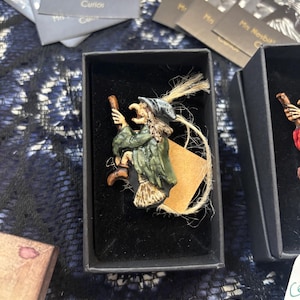 May include: A small, hand-painted witch figurine in a black box. The witch wears a green coat, a gray hat, and holds a broom. A tan tag is attached with twine. The box is surrounded by other boxes and cards that read "Mrs. Nesbit's Curious Curios."