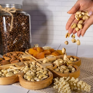 Könnte beinhalten: Ein Holztablett mit fünf Fächern, gefüllt mit Mandeln, Cashewnüssen, Pistazien und getrockneten Aprikosen. Eine Hand füllt das Tablett mit Pistazien.