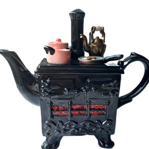 May include: A black ceramic teapot in the shape of a vintage stove, with a spout and handle. The stove top has miniature pots, a kettle, and a frying pan. The stove's front features red accents and decorative details.