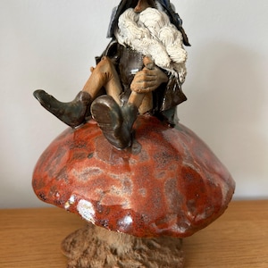 Puede incluir: Figura de gnomo de cerámica con una larga barba blanca y un gorro puntiagudo, sentado sobre una gran seta roja. El gnomo lleva un abrigo y botas marrones. La seta tiene una superficie texturizada y un tallo marrón claro.