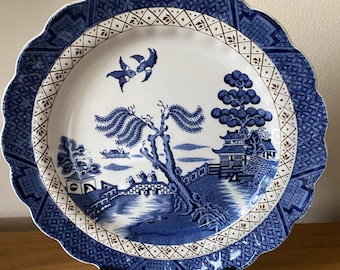 May include: A blue and white porcelain plate with a scalloped edge and a gold rim. The plate features a traditional willow pattern with a tree, a bridge, a pagoda, and birds.