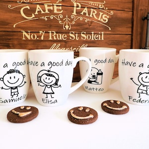 Puede incluir: Cuatro tazas de cerámica blanca con asas negras. Cada taza tiene una ilustración en blanco y negro de una persona con el texto "Have a good day" y un nombre en la parte inferior. Los nombres son Samuele, Elisa, Matteo y Federico. Hay tres galletas de chocolate con caras sonrientes.