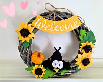 Welcome wreath featuring hand-crafted felt designs, including sunflowers, a pumpkin, and a cute blackbird. A perfect gift for your home.