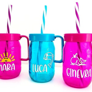 Peut inclure: Trois gobelets colorés de style bocal Mason avec poignées et pailles. Les gobelets sont roses et bleus, chacun avec un design et un nom différents : Mara, Luca et Ginevra. Les pailles sont rayées.