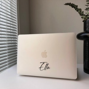May include: A white laptop computer with a black Apple logo and the name "Ella" written in black cursive on the lid.
