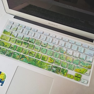 May include: A laptop keyboard cover with a green and yellow artistic design, resembling a painting. The cover is on a MacBook Air laptop. A sticker with the words "VAN GOGH" is on the laptop.
