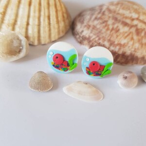 May include: A pair of colorful fishbowl earrings with a red fish and green seaweed inside. The earrings are made of polymer clay and have a white background with blue water.