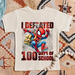 Puede incluir: Una camiseta blanca con una imagen de dibujos animados roja y azul de Spiderman con una mochila y el texto "I Defeated 100 Days of School".