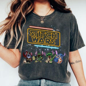 May include: A dark grey t-shirt featuring a graphic print of Stitch characters from the Disney movie Lilo & Stitch dressed as Star Wars characters. The text "Stitch Wars" is printed in yellow on a black rectangle with red and blue lightsabers.