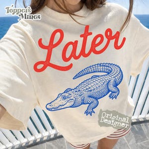 May include: Cream-colored t-shirt with the word "Later" in red script above a blue alligator illustration. The shirt also features the text "Original Designer" in a small gray box.