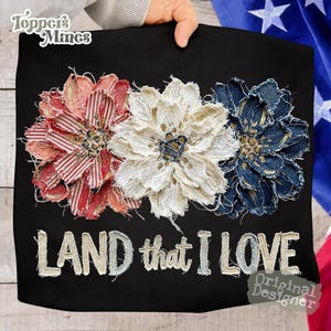May include: Black fabric with three fabric flowers in red, white, and blue, with the text "LAND that I LOVE". The flowers are made of layered fabric, with the red flower having a striped pattern. The white flower is made of lace, and the blue flower is made of denim.