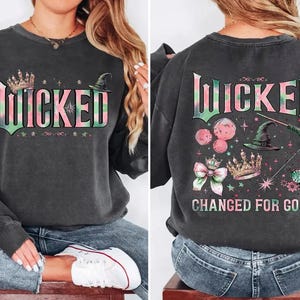 Könnte beinhalten: Ein schwarzer Pullover mit einem pinkfarbenen und grünen Grafikdruck, auf dem "Wicked Changed for Good" steht. Die Grafik enthält eine Krone, einen Hexenhut und andere fantasievolle Details.