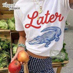 May include: White t-shirt with the word "Later" in red script and a blue alligator illustration. The shirt has a crew neck and short sleeves. The image also shows a person holding two apples.
